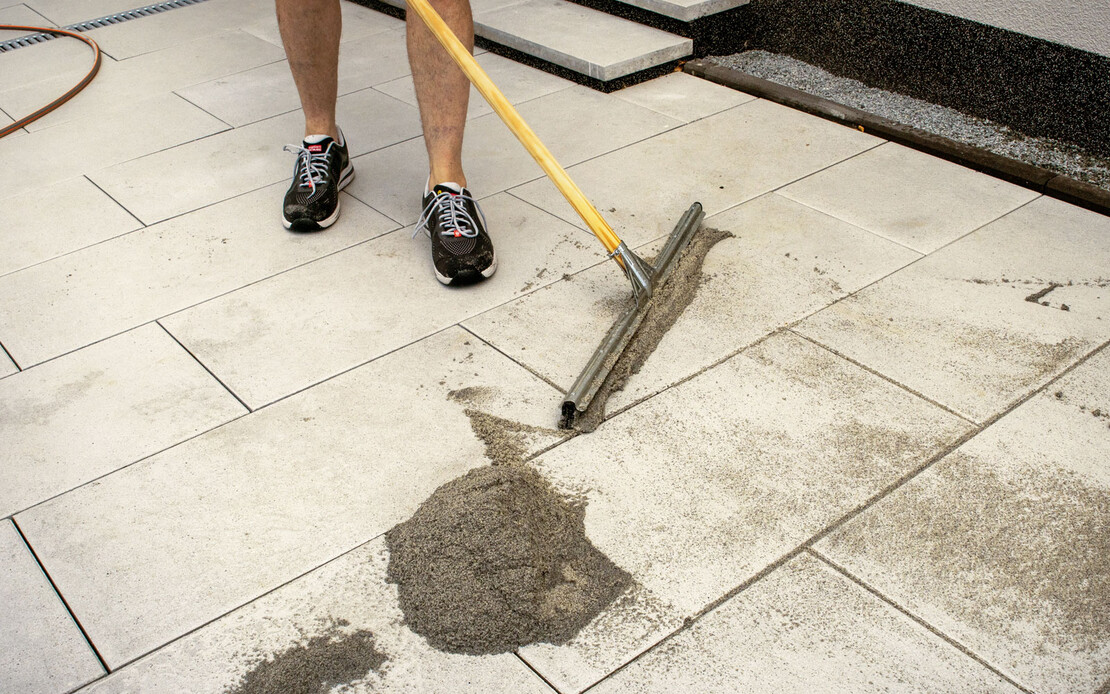 Der Pflasterfugenmörtel Fix von Baumit wird mit einem Gummischieber in die Fugen der Terrassenplatten verteilt.